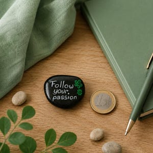 May include: A smooth, black, oval-shaped stone featuring the white hand-painted text "Follow your passion" and small green flower accents. A one-euro coin is placed next to the stone for scale, showing its size relative to the currency.