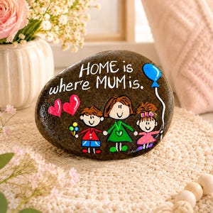 Peut inclure: Un galet brun foncé lisse peint avec une illustration de famille et le texte blanc "HOME is where MUM is." L'œuvre représente un parent et deux enfants, deux cœurs rouges et un ballon bleu, le tout réalisé avec de la peinture colorée.