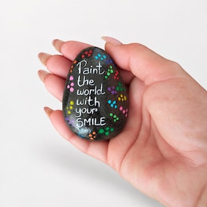 Peut inclure: Une pierre lisse, gris foncé, en forme d'œuf, avec le texte blanc peint à la main "Paint the world with your SMILE" entouré de petits points multicolores dans des tons de rouge, bleu, vert, jaune et violet.