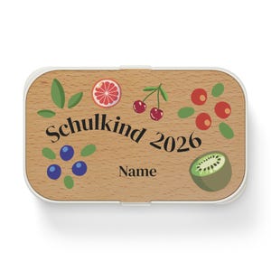 Op de afbeelding: Een rechthoekige lunchbox met een houten look deksel met fruitillustraties en de tekst "Schulkind 2026". Het ontwerp bevat een grapefruit schijf, kersen, bosbessen en een kiwi. De doos heeft een witte rand.
