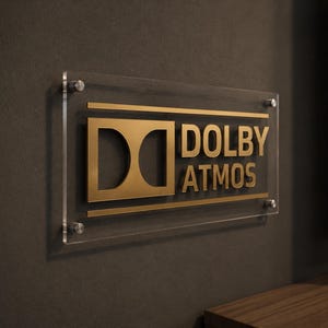 Może przedstawiać: Przezroczysty akrylowy znak z złotym napisem "DOLBY ATMOS" zamontowany na ciemnoszarej ścianie. Prostokątny znak z zaokrąglonymi rogami jest przymocowany czterema srebrnymi śrubami. Znak znajduje się nad drewnianą p&oacute;łką.