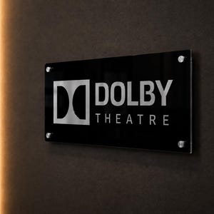 Könnte beinhalten: Ein rechteckiges schwarzes Schild mit glänzender Oberfläche, das das silberne Dolby-Logo und den Text DOLBY THEATRE in Großbuchstaben zeigt. Das Schild ist mit vier silbernen, runden Abstandshaltern an den Ecken an einer dunklen Wand befestigt.