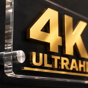 May include: A rectangular sign featuring the gold-colored text 4K ULTRAHD mounted on a clear acrylic panel. The sign is secured with silver-toned metal standoffs at the corners, creating a layered effect with the bold, metallic lettering.