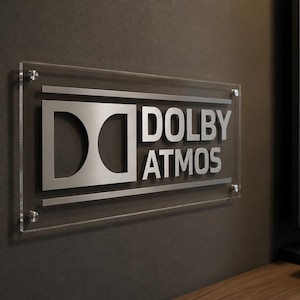 Może przedstawiać: Przezroczysty akrylowy znak z logo Dolby Atmos w kolorze srebrnym. Znak jest zamontowany na ciemnoszarej ścianie za pomocą srebrnych element&oacute;w mocujących. Logo zawiera symbol Dolby i słowa "DOLBY ATMOS".