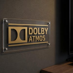 Może przedstawiać: Przezroczysty akrylowy znak z złotym logo "DOLBY ATMOS", zamontowany na ciemnoszarej ścianie. Prostokątny znak jest zabezpieczony czterema srebrnymi śrubami. Widoczny jest czarny soundbar i telewizor.