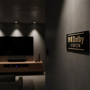 K&ouml;nnte beinhalten: Ein rechteckiges schwarzes Schild mit goldener Aufschrift Dolby Vision, montiert an einer dunkelgrauen, strukturierten Wand. Das Schild verf&uuml;gt &uuml;ber einen horizontalen goldenen Linienakzent und befindet sich in einem schwach beleuchteten Raum.