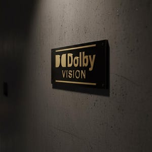 K&ouml;nnte beinhalten: Ein rechteckiges, schwarzes Wandschild mit dem goldfarbenen Dolby Vision-Logo und Schriftzug. Das Schild ist an einer dunklen, strukturierten Betonoberfl&auml;che befestigt, wobei die metallische Schrift deutlich auf dem mattschwarzen Hintergrund hervorsticht.