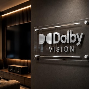 K&ouml;nnte beinhalten: Ein rechteckiges Wand-Schild aus klarem Acryl mit dem silbernen, metallischen Dolby Vision Logo und Schriftzug. Das Schild ist mit vier runden, silbernen Metallabstandshaltern an den Ecken an einer dunklen Wand befestigt, nahe einem Heimkinosystem.