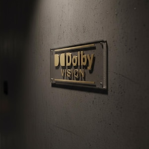 K&ouml;nnte beinhalten: Ein rechteckiges Wandschild mit dem goldenen Dolby Vision-Logo. Das Schild besteht aus einer transparenten Acrylplatte mit goldenen horizontalen Akzenten und erhabener goldener Schrift, montiert an einer dunkelgrauen, strukturierten Betonwand.