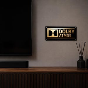 Może przedstawiać: Nowoczesny zestaw rozrywkowy z dużym czarnym telewizorem, soundbarem i znakiem Dolby Atmos. Znak jest czarny ze złotymi literami. Czarny wazon z trzciną i świecznik znajdują się na drewnianej szafce.