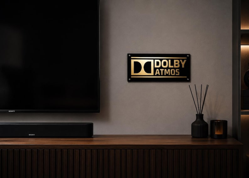 Może przedstawiać: Nowoczesny zestaw rozrywkowy z dużym czarnym telewizorem, soundbarem i znakiem Dolby Atmos. Znak jest czarny ze złotymi literami. Czarny wazon z trzciną i świecznik znajdują się na drewnianej szafce.
