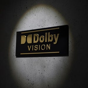 K&ouml;nnte beinhalten: Ein rechteckiges schwarzes Schild mit dem goldfarbenen Dolby Vision Logo und Schriftzug. Das Schild wird von einem kreisf&ouml;rmigen Scheinwerfer beleuchtet, der das metallische Finish der Buchstaben und die klaren Kanten der Platte hervorhebt.