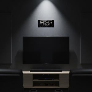 K&ouml;nnte beinhalten: Ein Heimkino-Setup mit einem gro&szlig;en Flachbildfernseher an einer dunkelgrauen Wand. &Uuml;ber dem Fernseher befindet sich ein rechteckiges schwarzes Schild mit dem wei&szlig;en Schriftzug Dolby VISION. Darunter steht ein helles Medienm&ouml;bel.