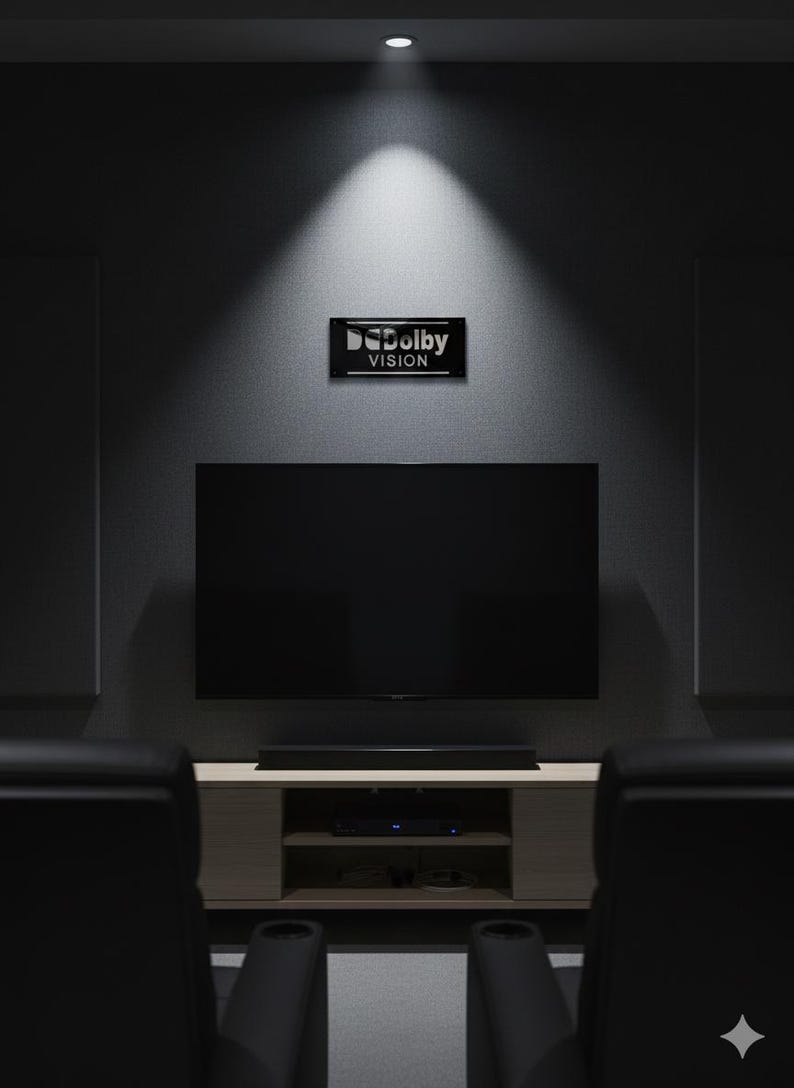 K&ouml;nnte beinhalten: Ein Heimkino-Setup mit einem gro&szlig;en Flachbildfernseher an einer dunkelgrauen Wand. &Uuml;ber dem Fernseher befindet sich ein rechteckiges schwarzes Schild mit dem wei&szlig;en Schriftzug Dolby VISION. Darunter steht ein helles Medienm&ouml;bel.