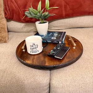 May include: A round, dark stained wooden serving tray with brass handles. The tray holds a white mug with the text "My Best Friend Has Four Paws", a small potted plant, a book, and two remote controls. The tray is on a beige couch.