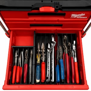 Op de afbeelding: Een rode Milwaukee Packout gereedschapskist met een open lade met zwarte kunststof verdelers. De lade is georganiseerd met diverse handgereedschappen, waaronder tangen en moersleutels met rode, blauwe en zilverkleurige handgrepen.