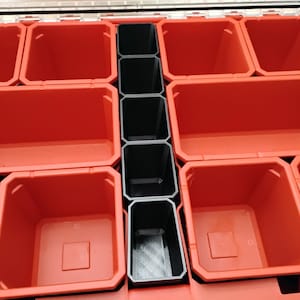 May include: A red plastic tool organizer case featuring multiple open compartments of varying sizes. A central column of five narrow black rectangular bins divides the larger red bins, providing organized storage for small hardware and tools.