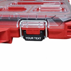 Może przedstawiać: Czerwona plastikowa walizka organizator z przezroczystą pokrywą i wieloma wewnętrznymi przegródkami. Walizka posiada metalowy mechanizm zatrzaskowy z czarną wypustką, na której nadrukowano biały napis YOUR TEXT na przedniej stronie.