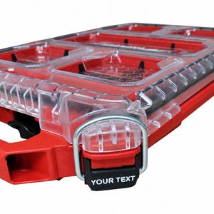 Puede incluir: Un estuche organizador de plástico rojo y transparente con múltiples compartimentos y una tapa transparente. El cierre frontal cuenta con una anilla en D de metal plateado y una pestaña negra con el texto blanco YOUR TEXT impreso.