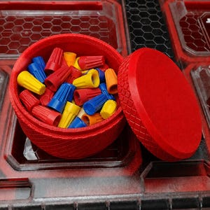 Può includere: Un contenitore rotondo rosso stampato in 3D con un esterno testurizzato a motivo di rombi, riempito con una collezione di piccoli connettori elettrici conici di vari colori, tra cui rosso, blu, giallo e arancione, con un coperchio rosso abbinato.