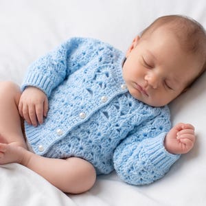 Könnte beinhalten: Eine hellblaue, langärmelige Häkeljacke mit strukturiertem Muschelmuster, gerippten Bündchen und vier kleinen, weißen, perlmuttartigen Knöpfen auf der Vorderseite, getragen von einem schlafenden Säugling auf einer weißen Unterlage.