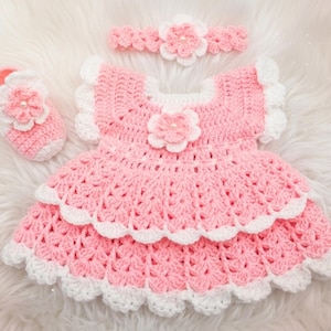 May include: A crochet set consisting of a pink and white dress with a layered skirt, a matching single shoe, and a headband. Each piece features a prominent pink flower with a central pearl-like bead and white scalloped trim edges.