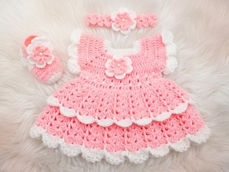 May include: A crochet set consisting of a pink and white dress with a layered skirt, a matching single shoe, and a headband. Each piece features a prominent pink flower with a central pearl-like bead and white scalloped trim edges.