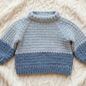 May include: A long-sleeved crochet jumper featuring a mock neck collar. The garment displays a colour-blocked design with a light grey upper section and a darker blue lower section, including matching blue cuffs and a ribbed hemline.