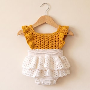 May include: A handmade baby romper in a mustard yellow and white crochet design. The top is mustard yellow with a ruffled edge, and the bottom is white with layered ruffles. The romper is hanging on a wooden hanger.