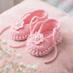 Puede incluir: Un par de zapatos de bebé tejidos a ganchillo en color rosa claro, con una flor decorativa en la punta, una perla blanca central en cada flor y una hilera de pequeñas perlas blancas a lo largo de la base con lazos de ganchillo a juego.