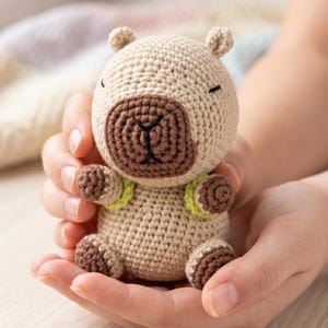 May include: A crocheted capybara plush toy in tan, brown, and green. The handmade stuffed animal has a round body, small ears, and a brown snout. The toy is held in open hands.