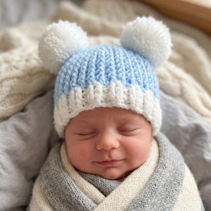 Puede incluir: Un gorro de bebé con un patrón de punto acanalado en azul claro y blanco con dos grandes pompones blancos en la parte superior. El gorro tiene un borde blanco doblado y está hecho de hilo suave, diseñado para ajustarse a la cabeza.