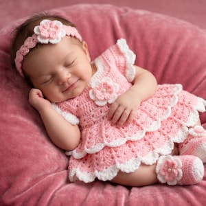 Pode incluir: Um vestido de crochê rosa claro e branco com faixa de cabelo e sapatinhos combinando. O vestido apresenta uma saia em camadas, mangas curtas e um detalhe floral no corpete. A faixa e os sapatinhos também incluem adornos florais.