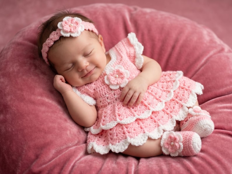 May include: A light pink and white crocheted dress with a matching headband and booties. The dress features a layered skirt, short sleeves, and a floral accent on the bodice. The headband and booties also include matching floral embellishments.