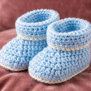 May include: A pair of light blue crochet booties featuring a thick, folded cuff and a white trim detail along the base and the top of the foot. The booties are constructed with a textured yarn stitch, creating a soft, rounded shape for the toe and heel.