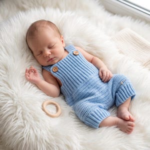 May include: A light blue knitted romper with ribbed texture and two small brown wooden buttons on the front straps. The garment features a simple design with cuffed leg openings, providing a soft and comfortable appearance for an infant.