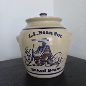 May include: Tan ceramic bean pot with a lid and two handles. The pot has blue trim and features the text "L.L. Bean Pot" and "Baked Beans". A blue and brown cabin graphic is also displayed on the pot.