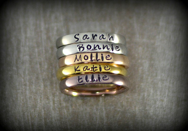 May include: Five stacked rings with different names engraved on them. The rings are silver, gold, and rose gold.