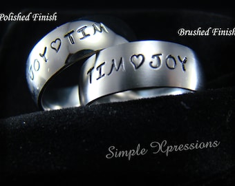 May include: Two silver rings with the words "Joy ♥ Tim" and "Tim ♥ Joy" engraved on them. The rings are on a black velvet surface. The words "Polished Finish" and "Brushed Finish" are written above the rings. The words "Simple Xpressions" are written below the rings.