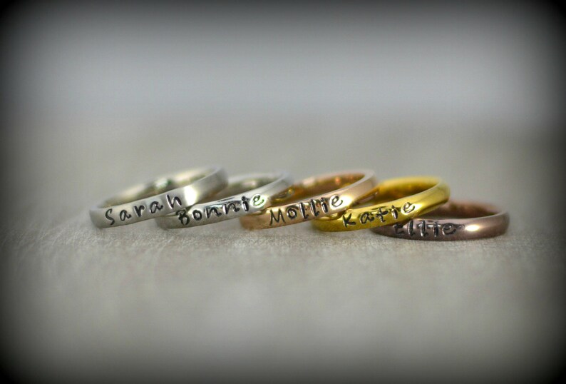 May include: Five silver, gold, and rose gold rings stacked on top of each other. Each ring has a name engraved on it: Sarah, Bonnie, Mollie, Katie, and Ellie.