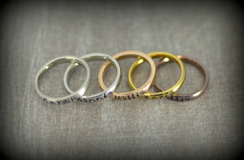 May include: Five silver, gold, and rose gold rings with names engraved on them. The names are Cora,  Naomi, Mollie, Ka-Flie, and Lillie.