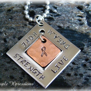 May include: Silver pendant necklace with a copper charm featuring a ribbon. The pendant is engraved with the words "Hope", "Strength", "Live", and "Dream".