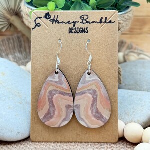 May include: A pair of teardrop-shaped earrings featuring a wavy, marbled pattern in shades of peach, tan, and muted purple. The earrings hang from silver-toned metal hooks and are displayed on a brown cardboard tag labeled Honey Bumble DESIGNS.