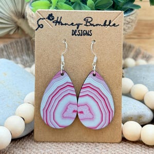 May include: Teardrop-shaped earrings featuring a pink and white agate-style pattern with silver-toned hooks. The earrings are displayed on a rectangular brown card labeled Honey Bumble DESIGNS in black ink, with a small bee icon next to the text.