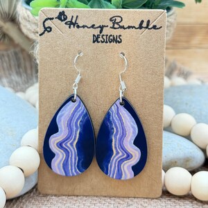 May include: Teardrop-shaped earrings featuring a dark navy blue background with a central wavy pattern of light purple and cream-colored lines. The earrings hang from silver-toned metal hooks and are displayed on a brown card labeled Honey Bumble DESIGNS.