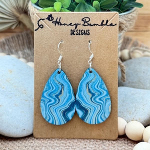 May include: Teardrop-shaped earrings featuring a blue, navy, and gold marbled pattern on a smooth surface. The earrings hang from silver-toned metal hooks and are displayed on a rectangular brown card labeled Honey Bumble DESIGNS.