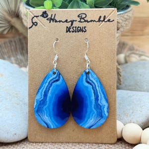 May include: Teardrop-shaped earrings featuring a vibrant blue agate stone pattern with concentric rings of dark and light blue hues. The earrings hang from silver-toned metal hooks and are displayed on a rectangular brown card labeled Honey Bumble DESIGNS.