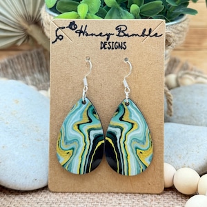May include: Teardrop-shaped earrings featuring a marbled pattern of teal, yellow, black, and white swirls. The earrings hang from silver-toned metal hooks and are displayed on a rectangular brown card labeled Honey Bumble DESIGNS.