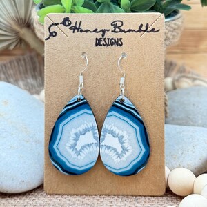 May include: Teardrop-shaped earrings featuring a blue and white agate-inspired pattern on a tan cardboard display card labeled Honey Bumble DESIGNS. The earrings have silver-toned metal hooks and a smooth, glossy finish with concentric rings.