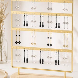 May include: A gold-toned metal earring display stand with a light wood base, featuring four horizontal bars with multiple holes. The stand holds various pairs of dangling earrings with silver, gold, black, and white beads in floral-like patterns.